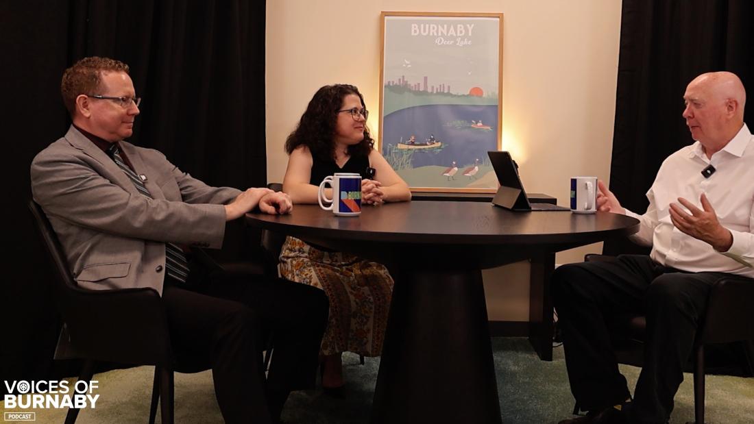 Three people seated around a round table during a podcast recording. Two mugs and a tablet sit on the table, with a Burnaby Deer Lake poster displayed on the wall behind them. The “Voices of Burnaby Podcast” logo appears in the bottom left corner.