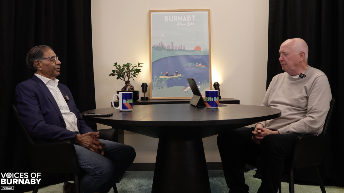 Two people sit across a round table in a podcast studio with Burnaby mugs, a tablet, and a framed Deer Lake artwork in the background. The Voices of Burnaby Podcast logo appears in the corner.