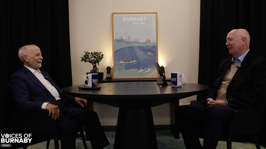 Two people seated across from each other at a round table in a podcast studio, with microphones, Burnaby mugs, and a framed Deer Lake poster in the background. The Voices of Burnaby Podcast logo appears in the bottom left corner.